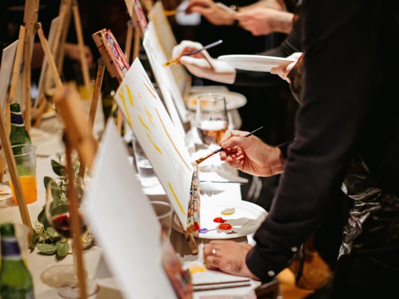 Canvases lined up on a table interspersed with wine glasses